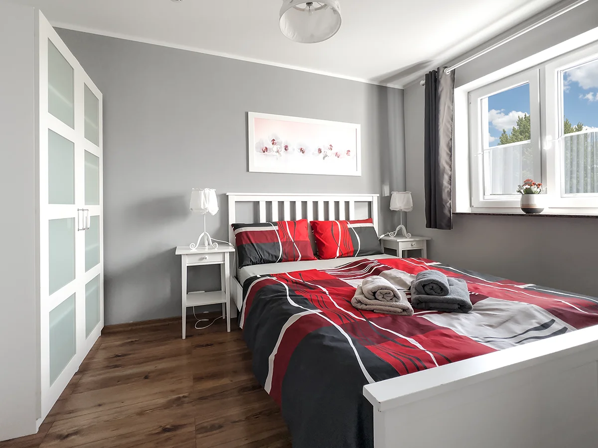 Modernes Schlafzimmer der Wohnung 4 mit weißem Doppelbett, rot-schwarzer Bettwäsche und Laminatboden.