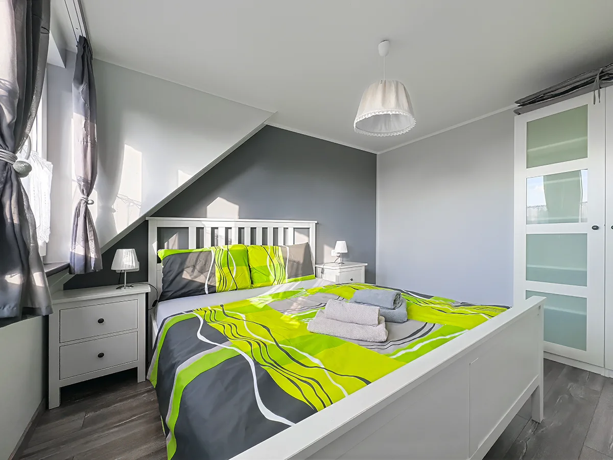 Schlafzimmer im Dachgeschoss der Wohnung 5 mit weißem Doppelbett, Dachschräge und moderner Bettwäsche in Grün-Grau.