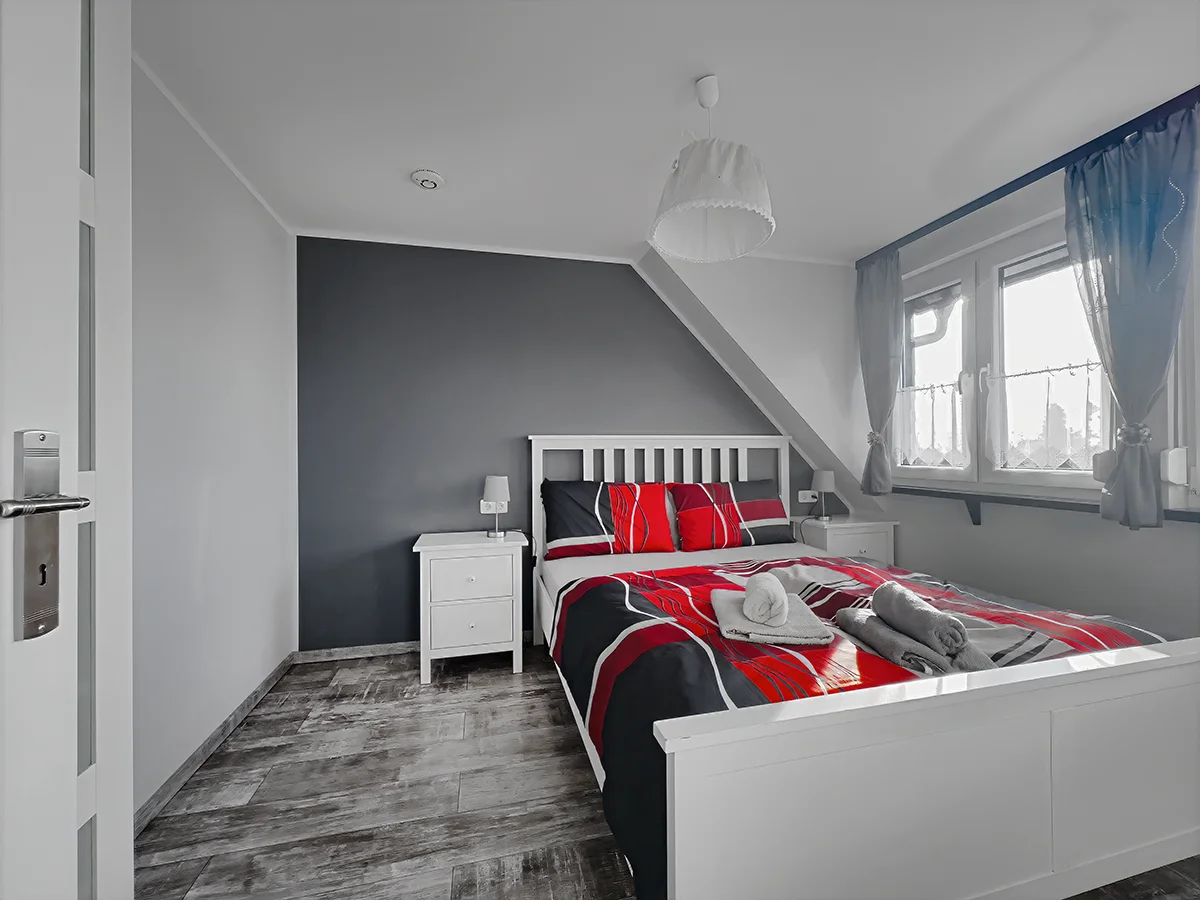Gemütliches Dachgeschoss-Schlafzimmer der Wohnung 6 mit Doppelbett, rot-schwarzer Bettwäsche und grauer Akzentwand.