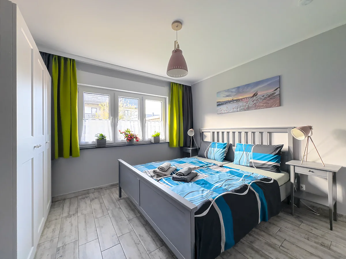 Schlafzimmer der Wohnung 8 mit massivem Holzbett, grüner Bettwäsche und zwei Fenstern für ein helles Ambiente.