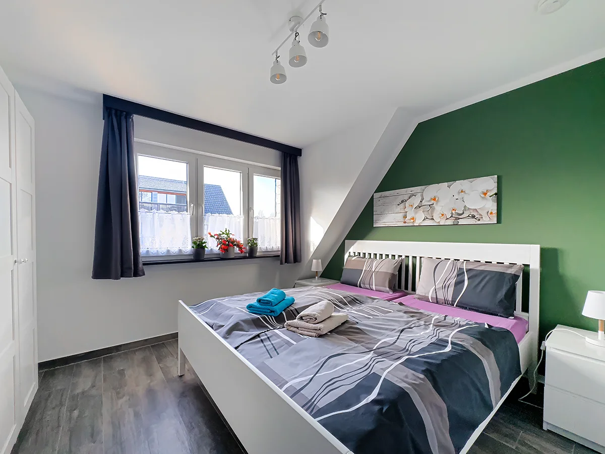Schlafzimmer im Dachgeschoss der Wohnung 10 mit weißem Doppelbett vor einer dunkelgrünen Akzentwand.