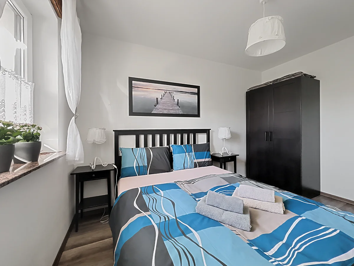 Gemütliches Schlafzimmer der Wohnung 3 mit Doppelbett, blauer Bettwäsche und Nachttischen unter einer dunklen Balkendecke.