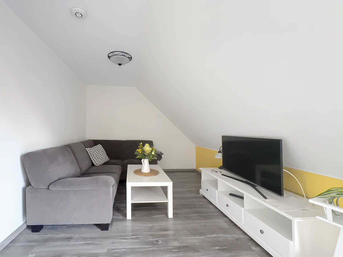 Gemütlicher Wohnbereich der Wohnung 5 mit grauem Ecksofa, weißem Couchtisch und Flachbild-TV unter der Dachschräge.